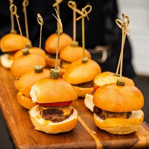 Miniature cheeseburger sliders with pickles on skewers, served on a wooden board.