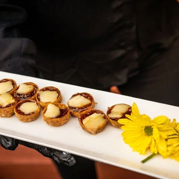 A server in black gloves holds a white tray of mini tarts with melted cheese and yellow flowers.
