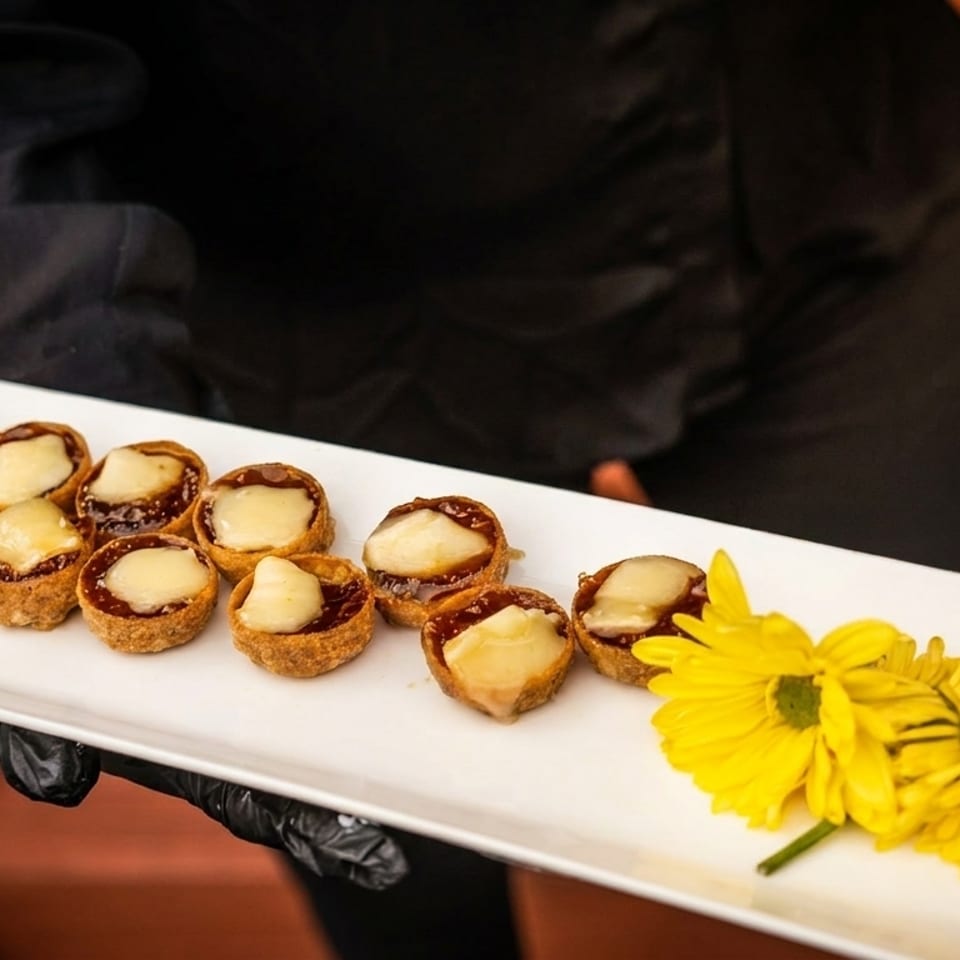 A server in black gloves holds a white tray of mini tarts with melted cheese and yellow flowers.